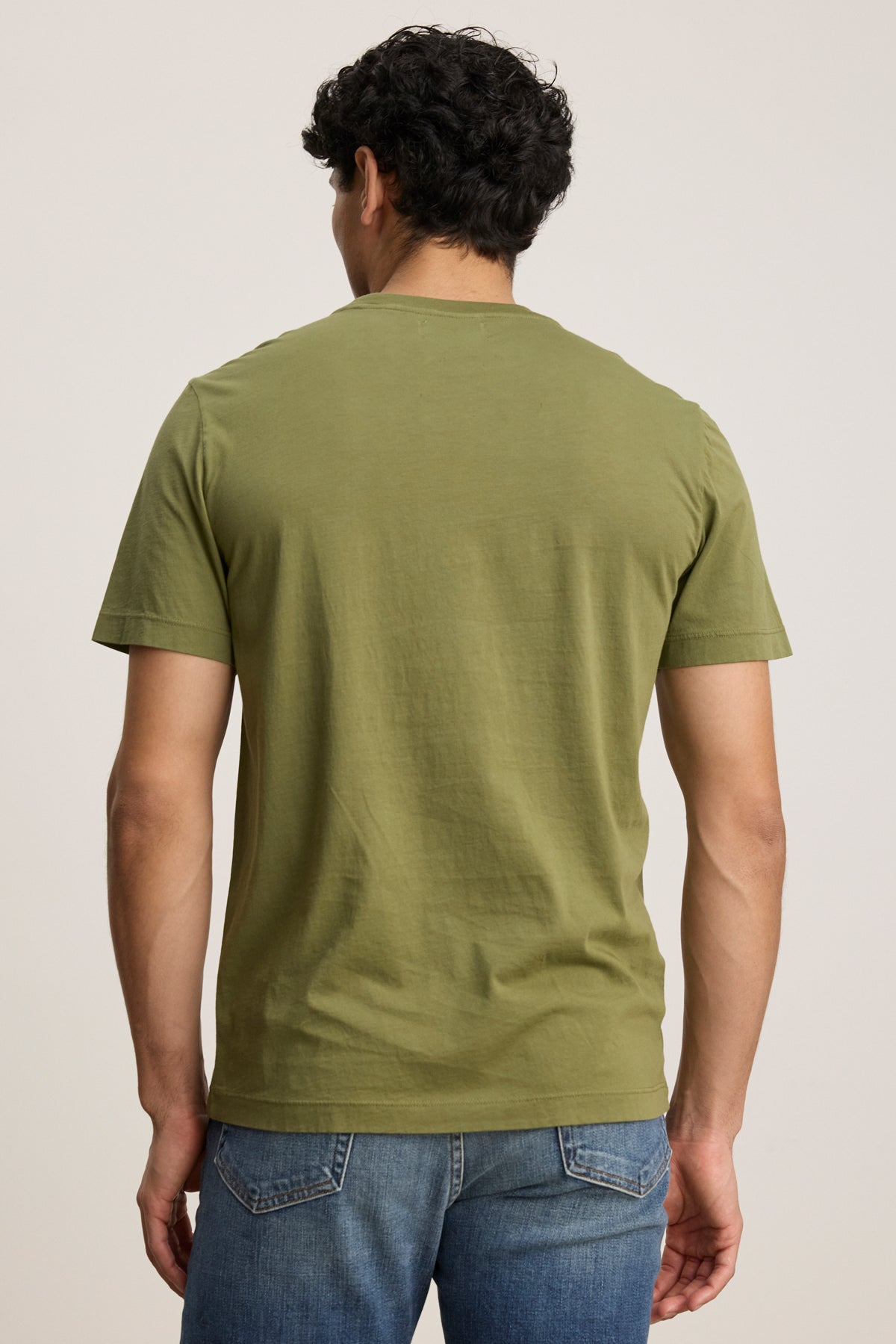 A man stands with his back to the camera, wearing the olive green HOWARD TEE by Velvet by Graham & Spencer, a lightweight Pima jersey short-sleeve shirt with a vintage feel, paired with blue jeans against a plain background.-39446027829441
