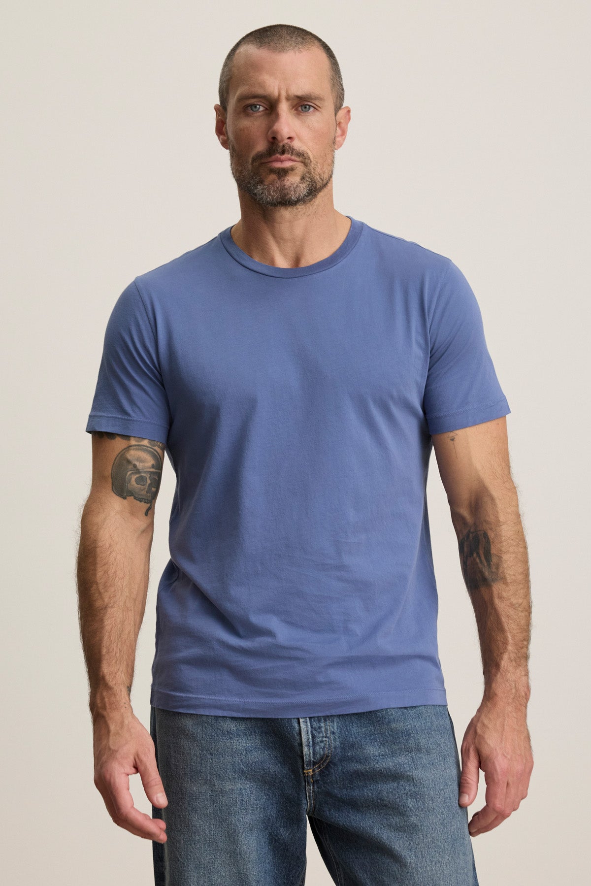 A man with a short beard and arm tattoos wears the HOWARD TEE by Velvet by Graham & Spencer in blue, paired with blue jeans, standing against a plain light background.-40606036230337