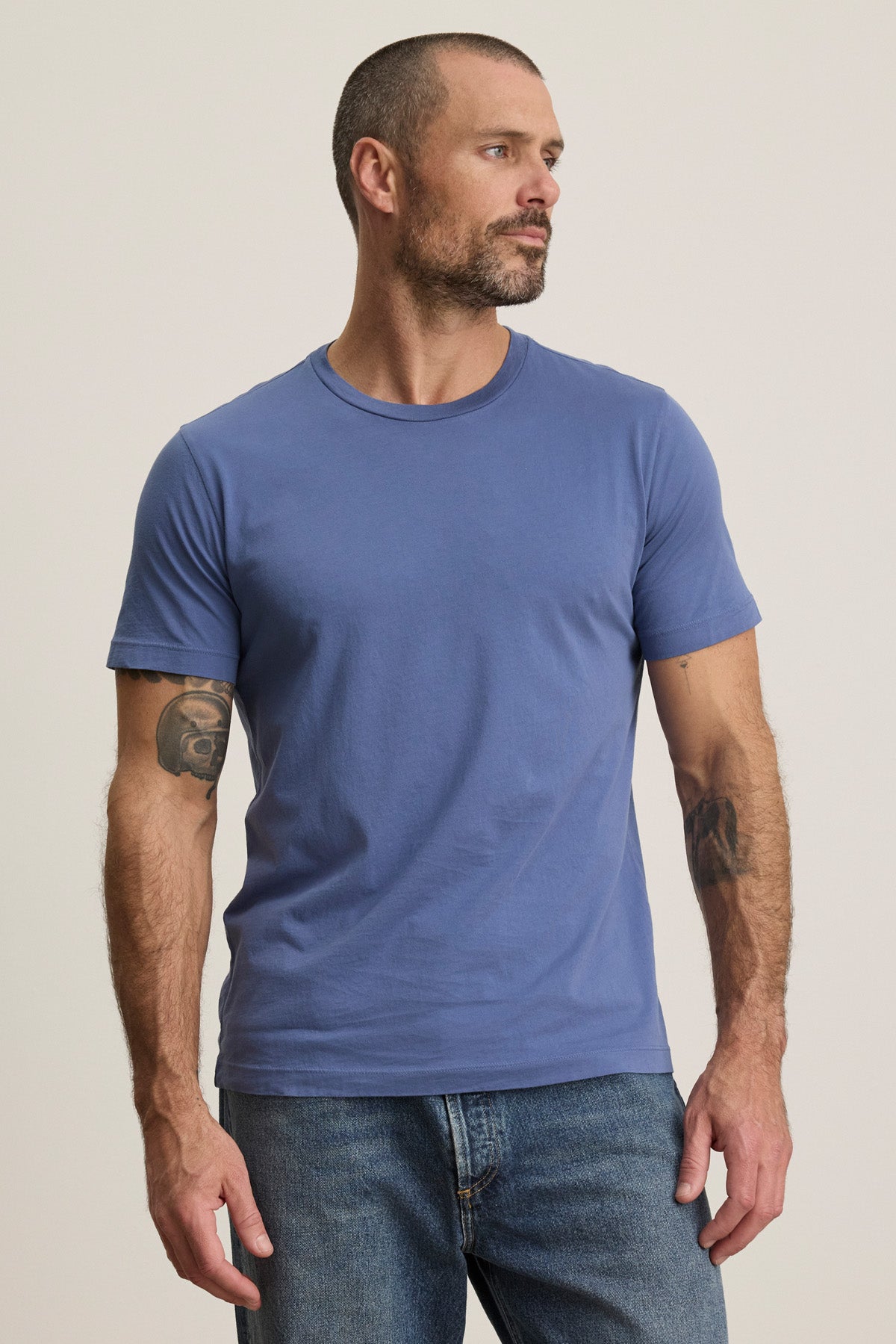 A man with short hair and tattooed arms is wearing the blue HOWARD TEE by Velvet by Graham & Spencer with jeans, standing against a plain light background.-40606036197569
