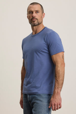 A man with short hair and a beard wears the HOWARD TEE by Velvet by Graham & Spencer, made from lightweight Pima jersey in blue, and jeans, standing against a plain light background, facing slightly to the side.