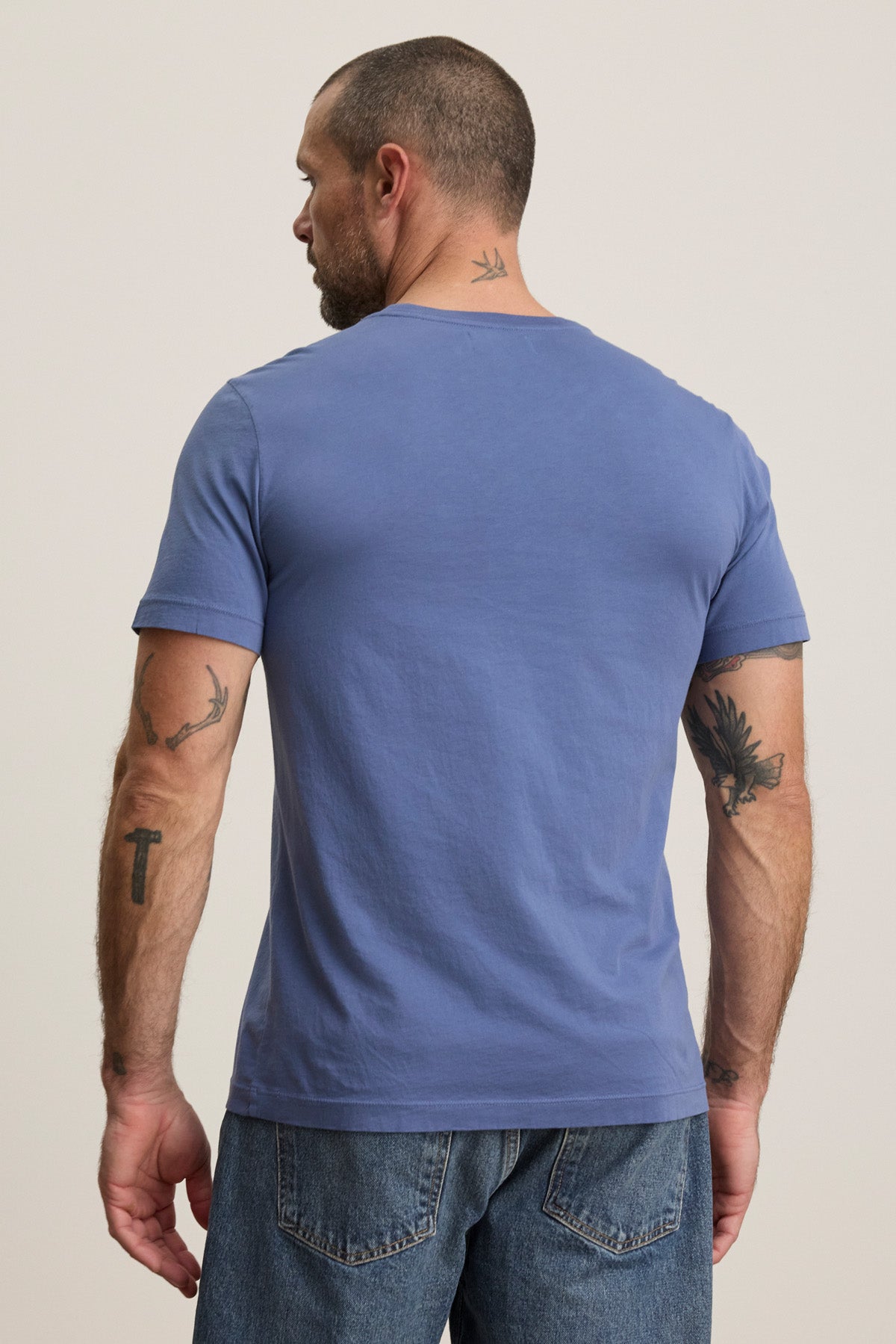 A man in the Velvet by Graham & Spencer HOWARD TEE, a blue lightweight Pima jersey t-shirt, and jeans stands with his back to the camera, revealing several tattoos on his arms and neck.-40606036132033
