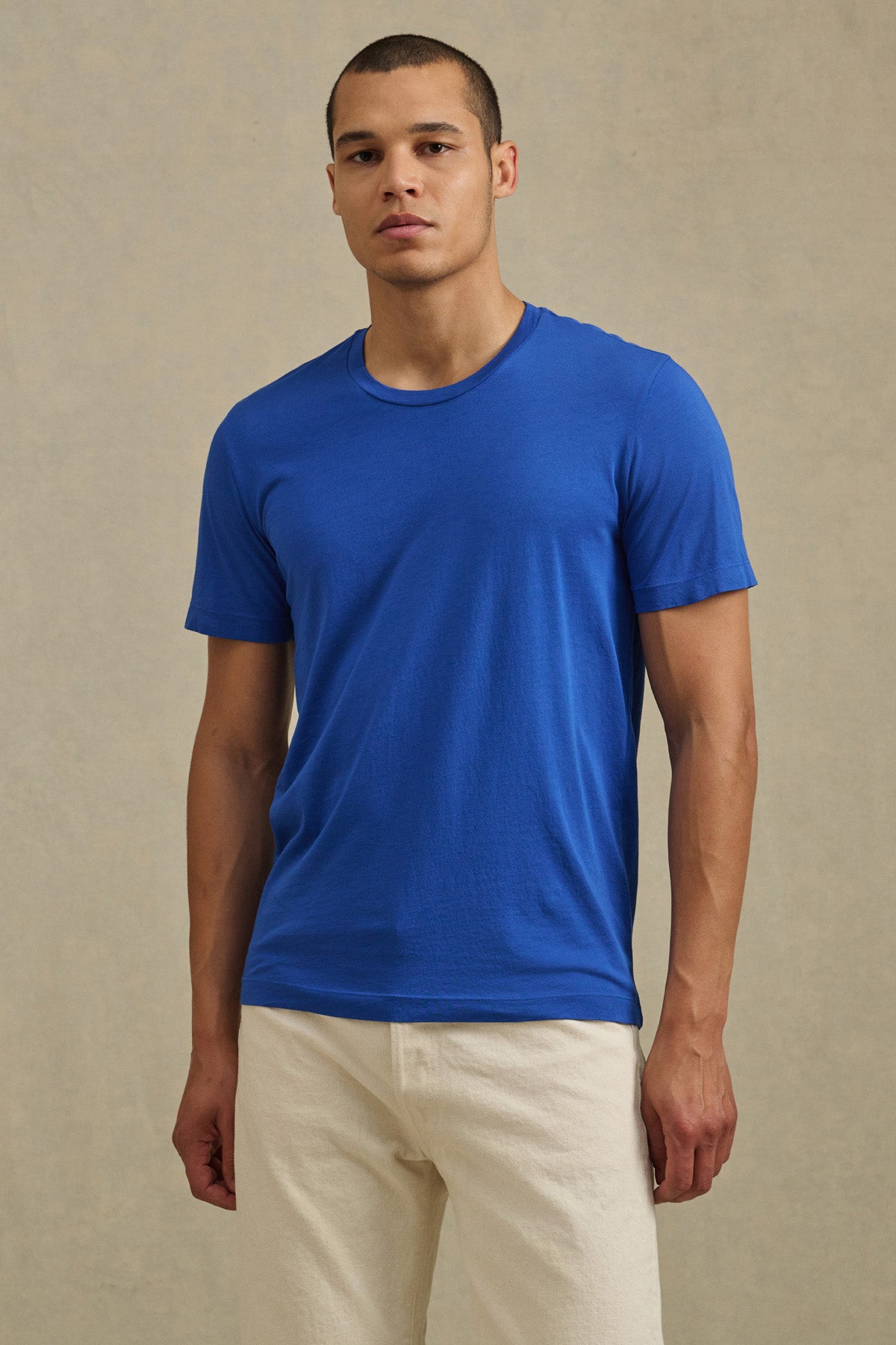 A man stands against a plain background wearing the HOWARD TEE by Velvet by Graham & Spencer, a blue short-sleeve Pima jersey shirt with a vintage feel and perfect fit, paired with light-colored pants.-41104752476353