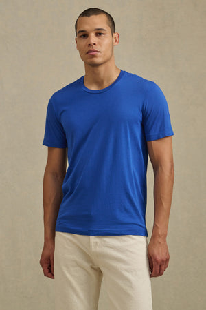 A man stands against a plain background wearing the HOWARD TEE by Velvet by Graham & Spencer, a blue short-sleeve Pima jersey shirt with a vintage feel and perfect fit, paired with light-colored pants.