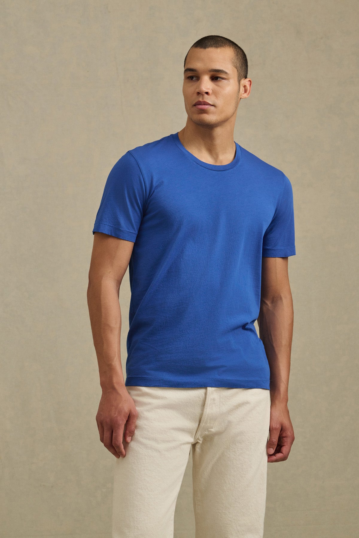 A man stands against a neutral background, looking slightly to the side for a vintage feel, wearing the HOWARD TEE from Velvet by Graham & Spencer in plain blue and off-white pants.-41104752443585