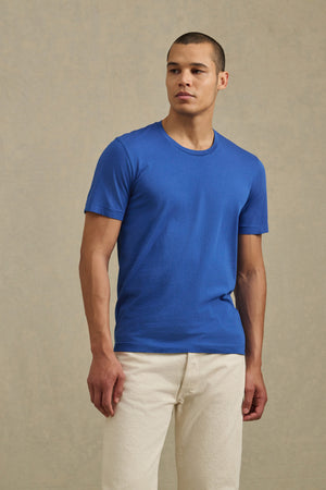 A man stands against a neutral background, looking slightly to the side for a vintage feel, wearing the HOWARD TEE from Velvet by Graham & Spencer in plain blue and off-white pants.