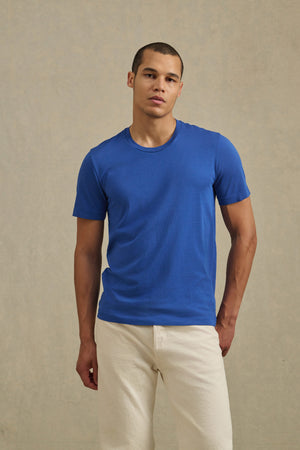 A person stands against a beige background, wearing the HOWARD TEE by Velvet by Graham & Spencer in blue and off-white pants, looking directly at the camera for that perfect fit.