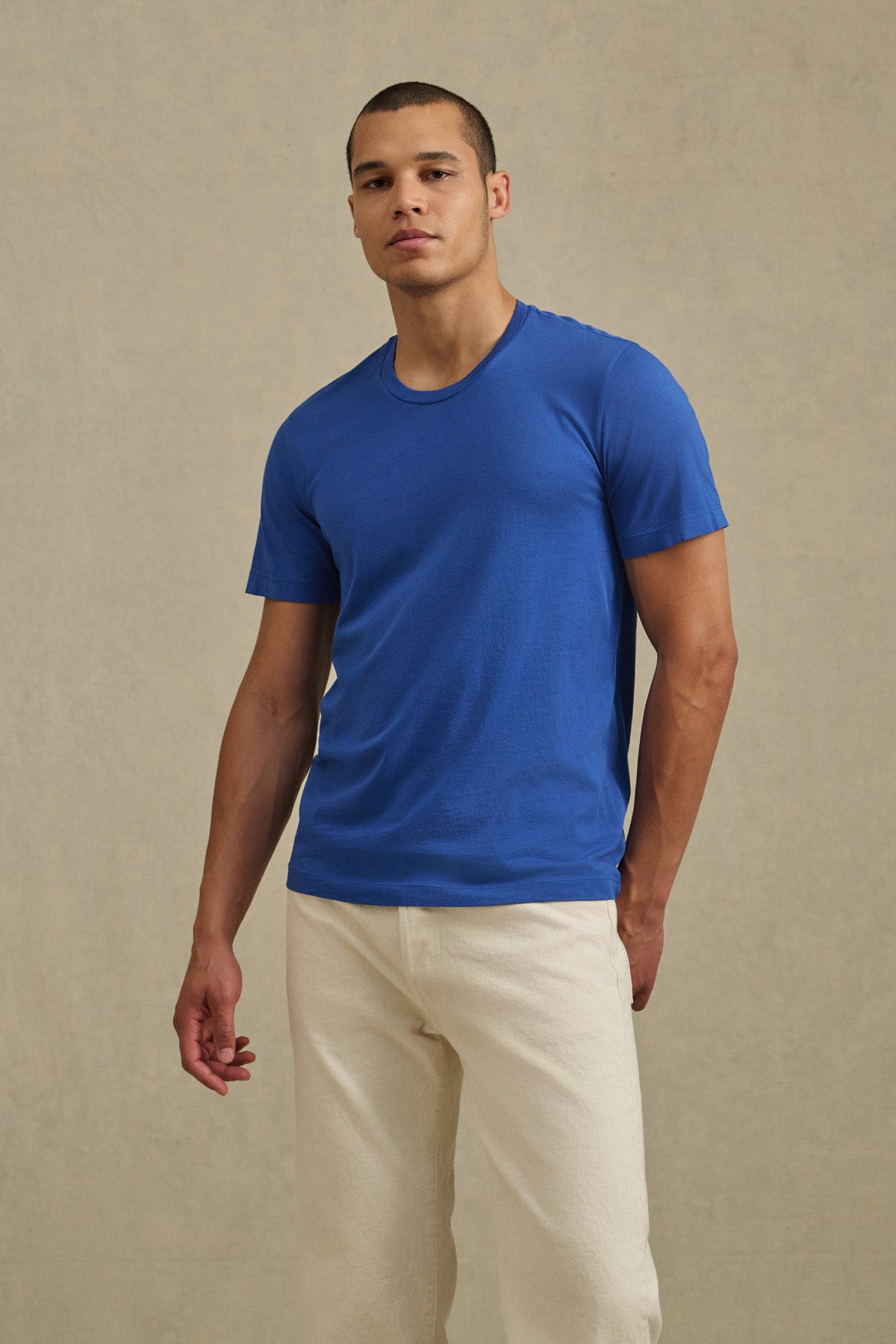 A person stands against a plain background wearing the HOWARD TEE by Velvet by Graham & Spencer in blue with off-white pants, showcasing a perfect fit.-41104752378049
