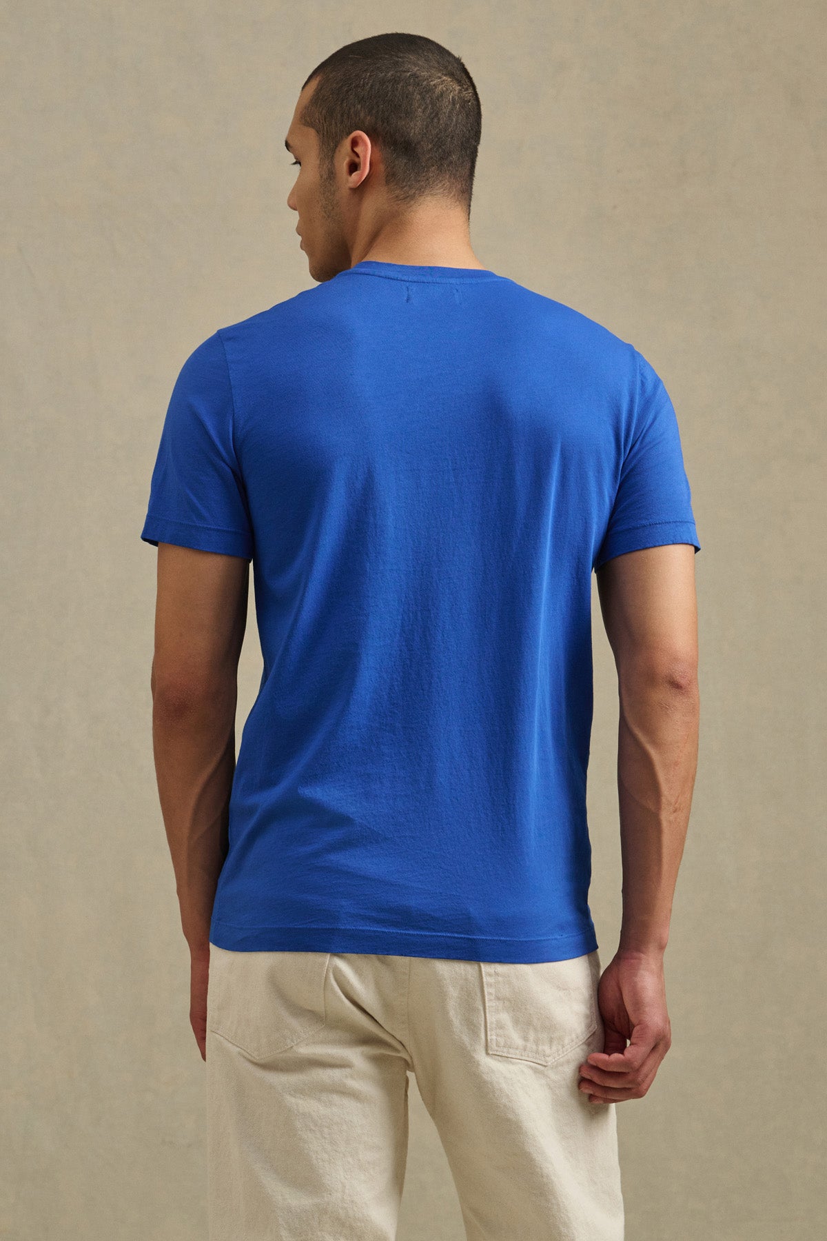 A person stands with their back to the camera, wearing the bright blue HOWARD TEE by Velvet by Graham & Spencer and light-colored pants against a plain, neutral background.