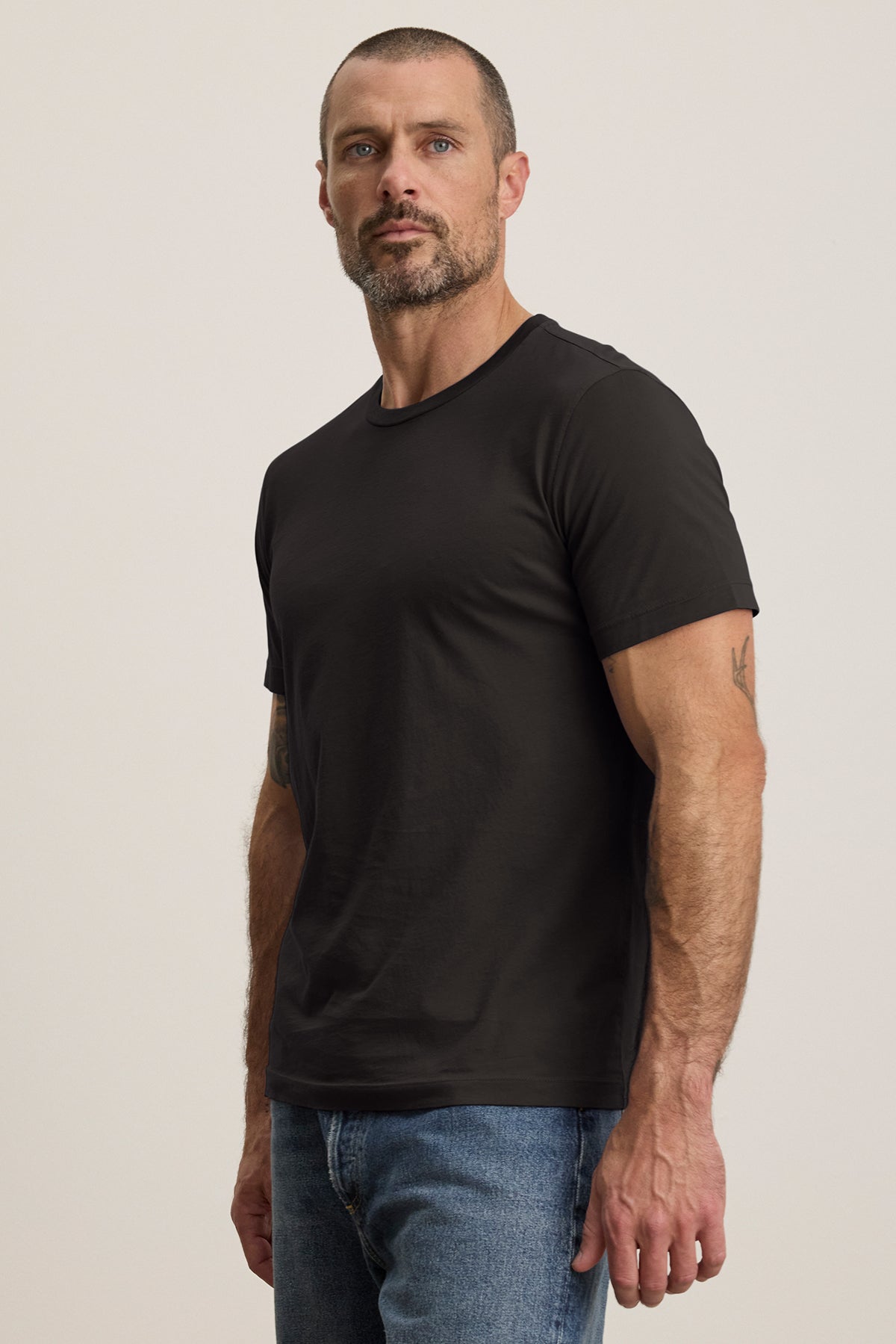   A bearded man in a black Velvet by Graham & Spencer HOWARD TEE, paired with blue jeans, faces the camera against a plain background. The tee is made from lightweight Pima jersey for comfort and an ideal fit. 