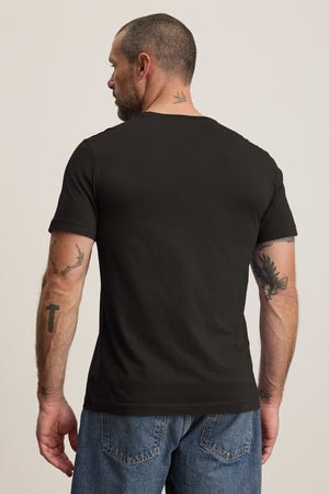 A tattooed man faces away, wearing the HOWARD TEE by Velvet by Graham & Spencer, a black t-shirt made from lightweight Pima jersey that offers a perfect fit, paired with blue jeans.