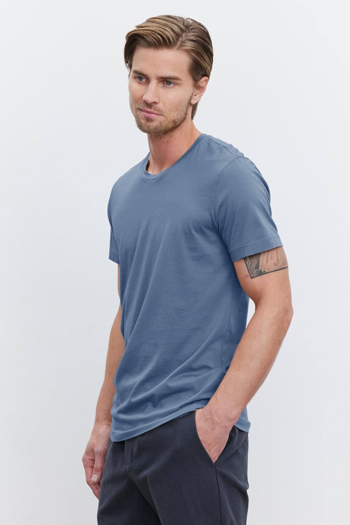   A man with short brown hair is standing with his right hand in his pocket, wearing a blue HOWARD TEE from Velvet by Graham & Spencer and dark pants that offer a perfect fit, against a plain white background. 