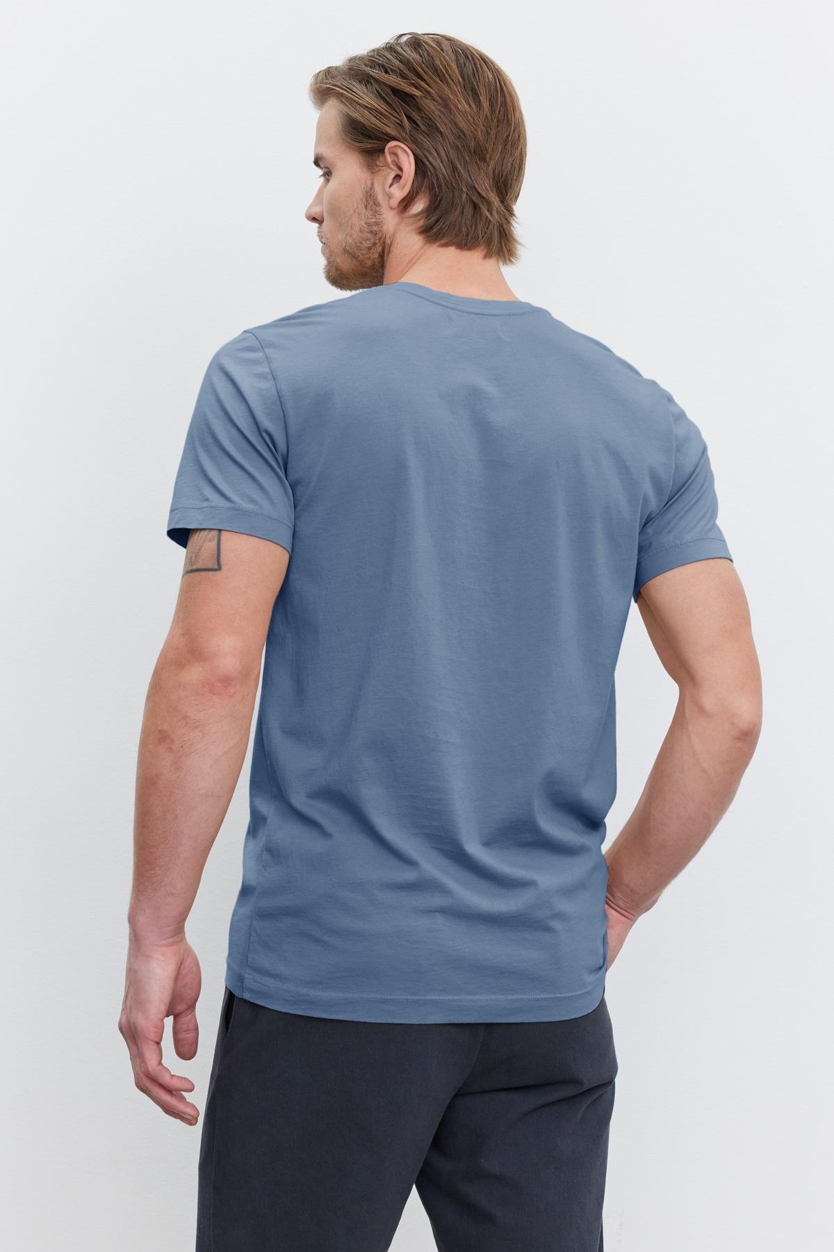   A man with light brown hair is standing with his back to the camera, wearing a perfectly fitted HOWARD TEE from Velvet by Graham & Spencer, made of soft Pima cotton jersey in a striking blue color, paired with dark pants for a vintage feel. 