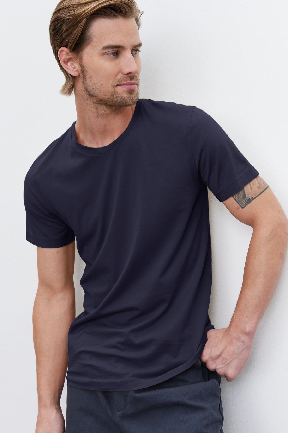 A man in a navy Howard Whisper Classic Crew Neck Tee by Velvet by Graham & Spencer with a vintage-feel softness leaning against a wall.-36273879417025