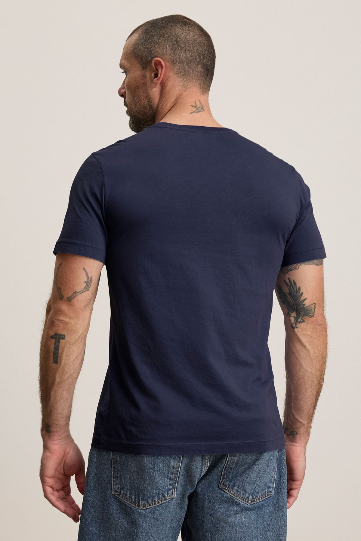 A man with short hair and tattoos stands with his back to the camera, wearing a navy HOWARD TEE by Velvet by Graham & Spencer in lightweight Pima jersey, paired with blue jeans.-39226005717185
