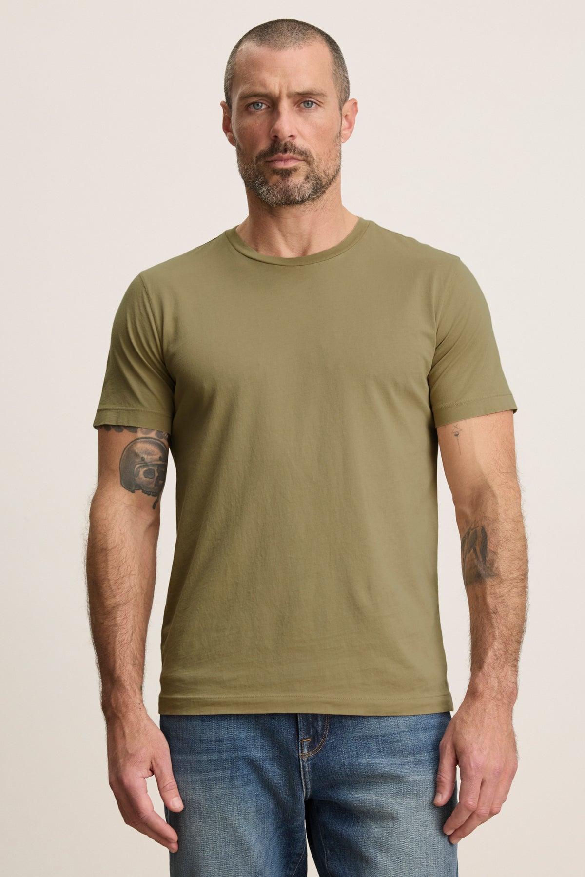 A man with short hair and a beard wears the olive green HOWARD TEE by Velvet by Graham & Spencer with blue jeans, standing against a plain background.-39680488472769