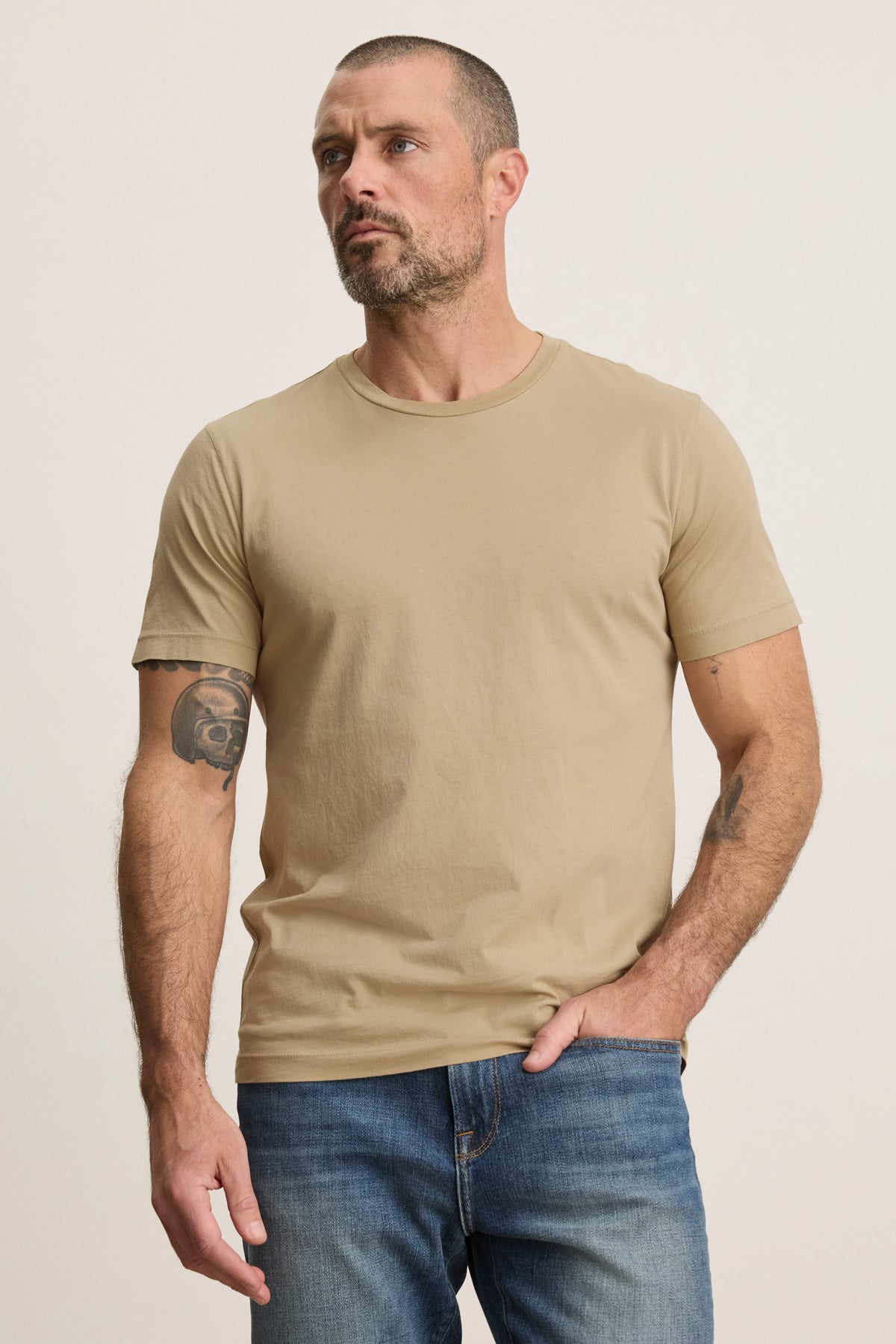 A man with short hair and a beard wears the beige HOWARD TEE by Velvet by Graham & Spencer with blue jeans, standing against a light background. A tattoo on his upper left arm adds vintage flair to this classic look.-39543512367297