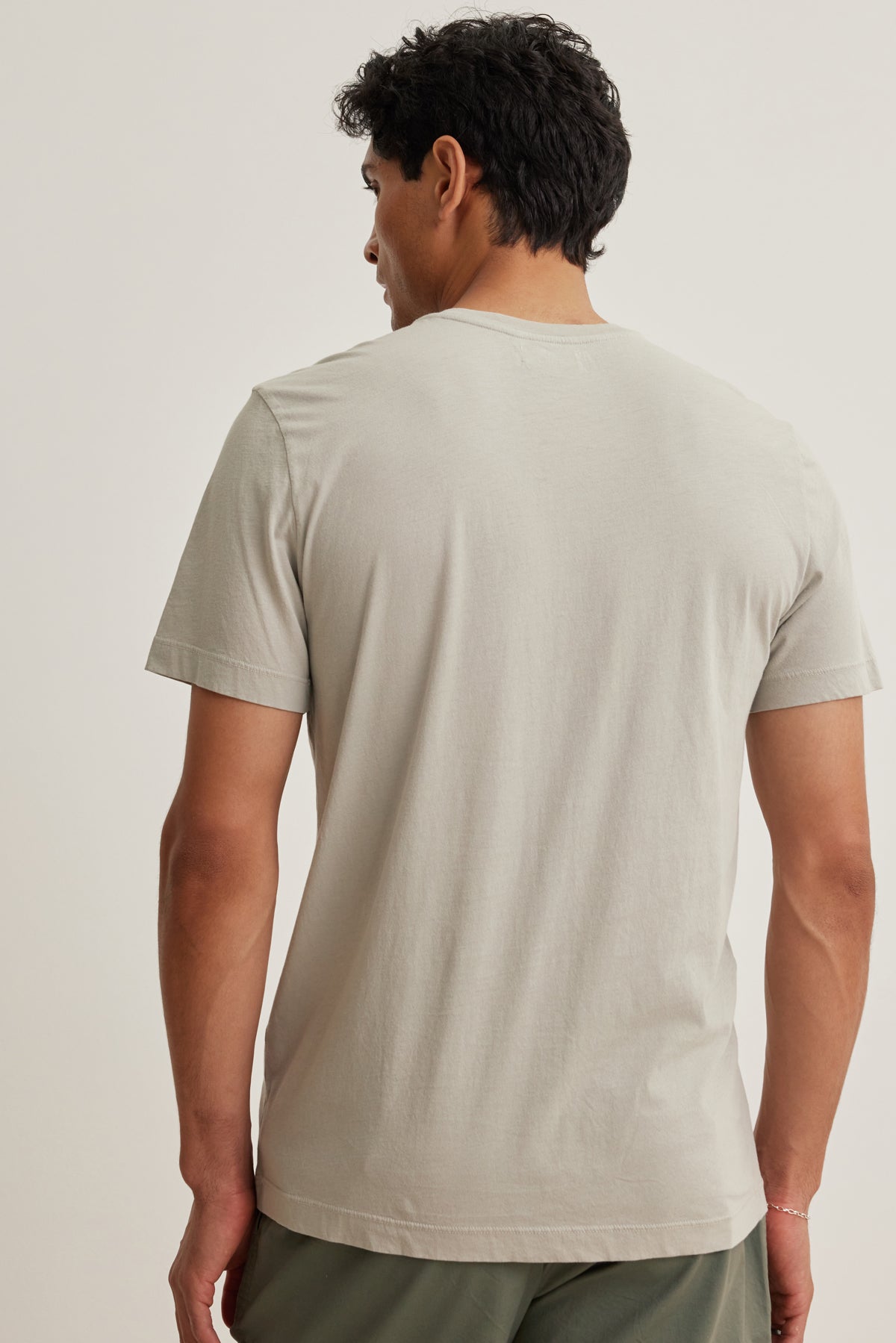 A man faces away from the camera wearing the HOWARD TEE by Velvet by Graham & Spencer, a light beige short-sleeve t-shirt made from lightweight Pima jersey, paired with green pants against a plain background.-39152696983745