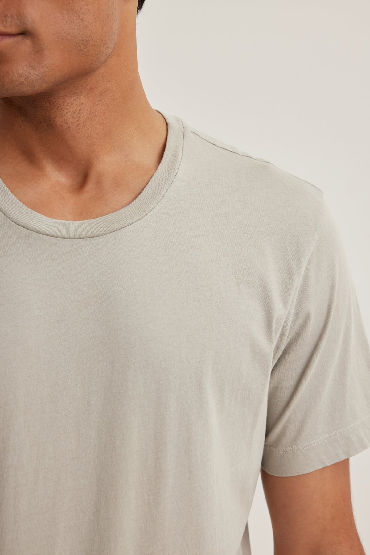 A person wearing the HOWARD TEE by Velvet by Graham & Spencer—a plain, light beige short-sleeve t-shirt in lightweight Pima jersey—is shown from shoulders to mid-torso, facing left against a neutral background.-39152696950977