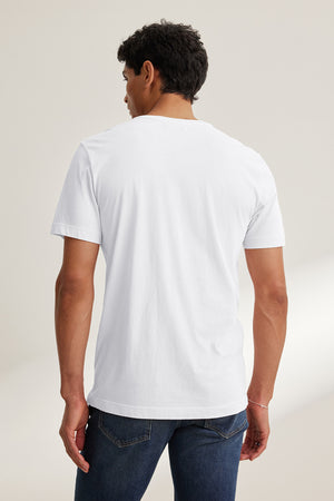 A man with short dark hair, facing away from the camera, wears Velvet by Graham & Spencer's HOWARD TEE made from lightweight Pima jersey and vintage-style blue jeans, creating a perfect fit in a neutral setting.