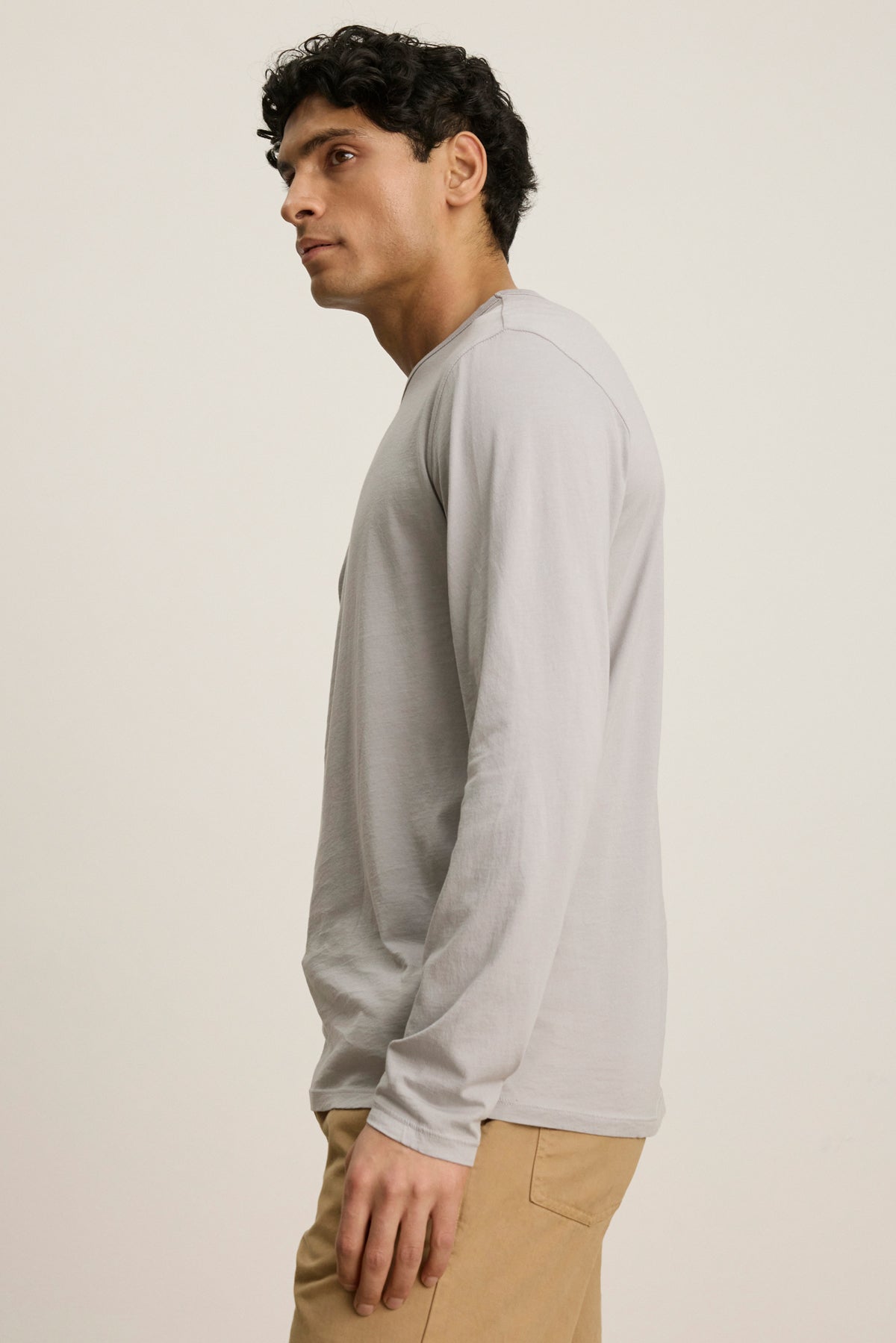   A man with short dark hair wears the Velvet by Graham & Spencer ROWLAND TEE—a light gray, long-sleeve crewneck—paired with tan pants, standing sideways against a plain light background. 