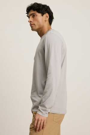 A man with short dark hair wears the Velvet by Graham & Spencer ROWLAND TEE—a light gray, long-sleeve crewneck—paired with tan pants, standing sideways against a plain light background.