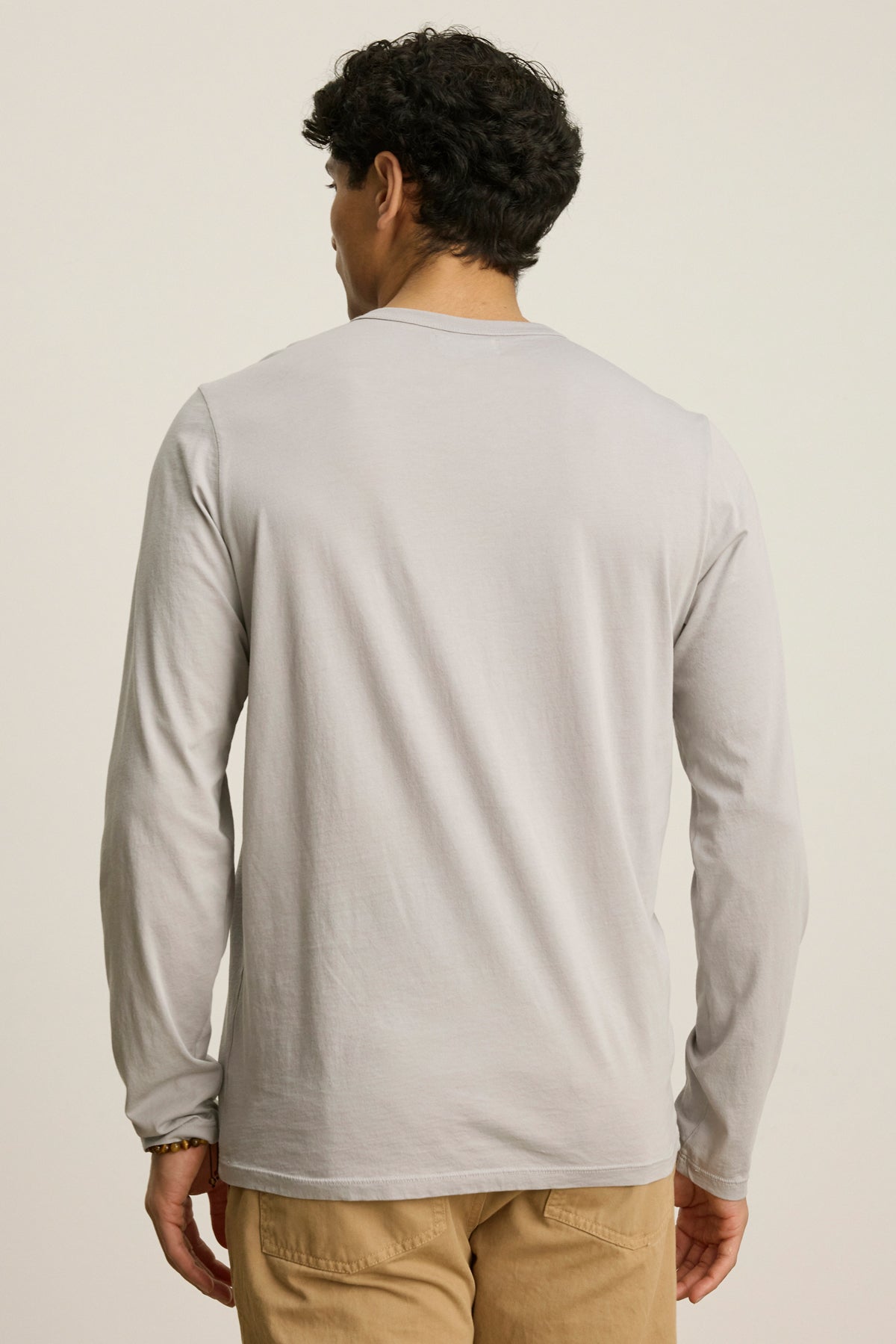 A man stands facing away, wearing the ROWLAND TEE by Velvet by Graham & Spencer—a light gray Pima jersey long-sleeve shirt with a crew neckline—paired with beige pants against a plain background.-39841089421505