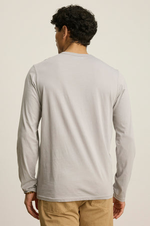 A man stands facing away, wearing the ROWLAND TEE by Velvet by Graham & Spencer—a light gray Pima jersey long-sleeve shirt with a crew neckline—paired with beige pants against a plain background.