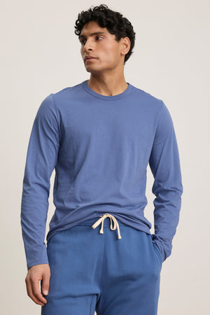 A man wearing the ROWLAND TEE by Velvet by Graham & Spencer and blue drawstring pants stands against a plain light background, looking to the side.
