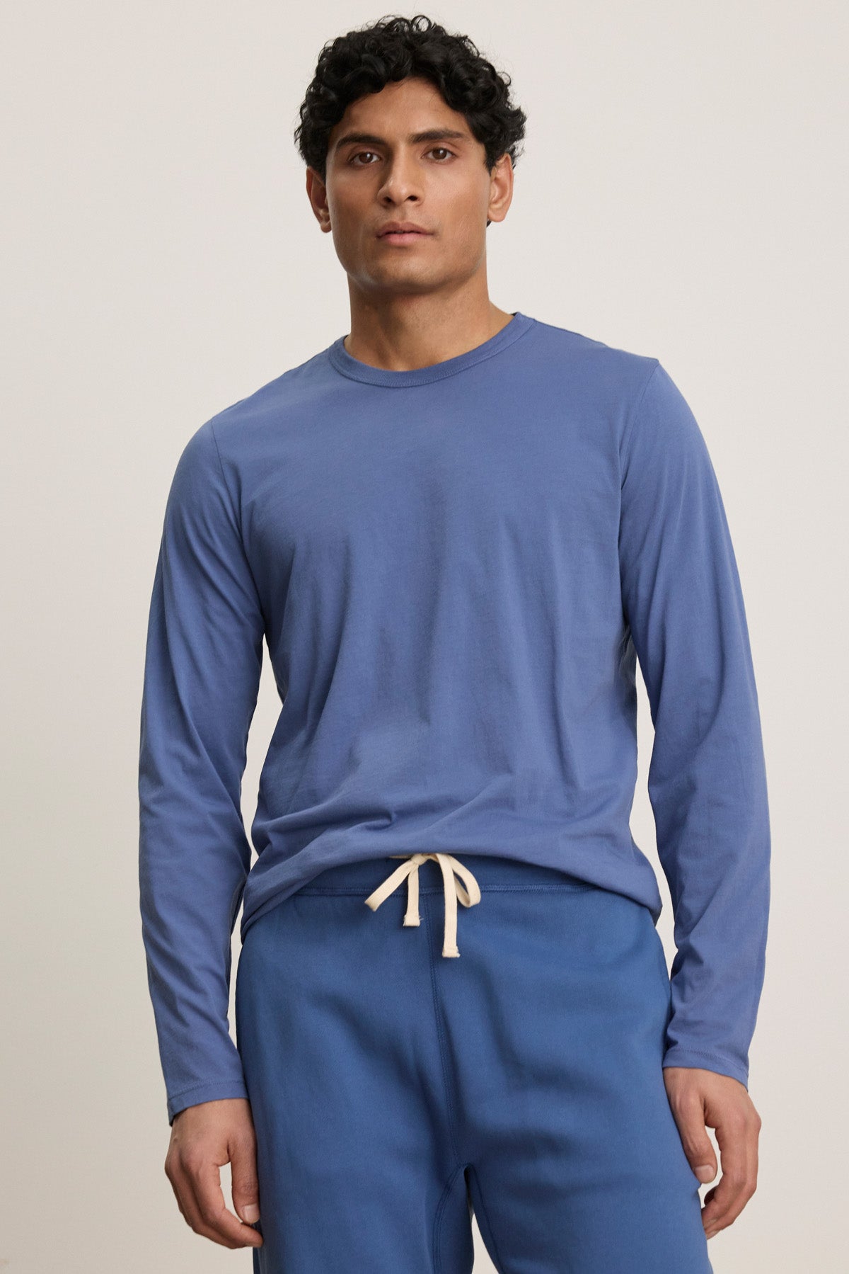 A man wears the ROWLAND TEE by Velvet by Graham & Spencer, a long-sleeve blue Pima jersey crewneck, with matching blue drawstring pants, standing facing the camera against a plain background.-40606036951233
