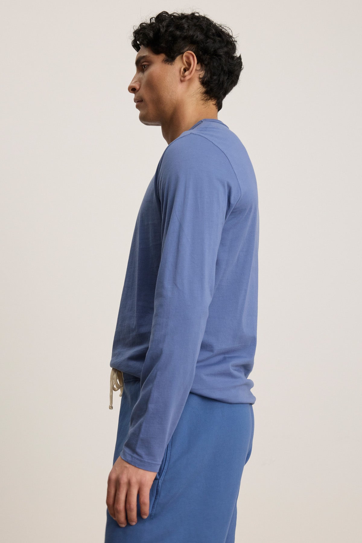 A person stands in profile against a plain, light background, wearing Velvet by Graham & Spencer's ROWLAND TEE—a long-sleeve Pima jersey crewneck—paired with blue pants.-40606036918465
