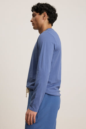 A person stands in profile against a plain, light background, wearing Velvet by Graham & Spencer's ROWLAND TEE—a long-sleeve Pima jersey crewneck—paired with blue pants.