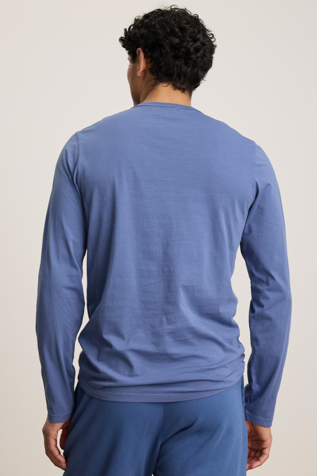 A person with short, curly hair stands with their back to the camera, wearing a blue ROWLAND TEE by Velvet by Graham & Spencer and matching blue pants.