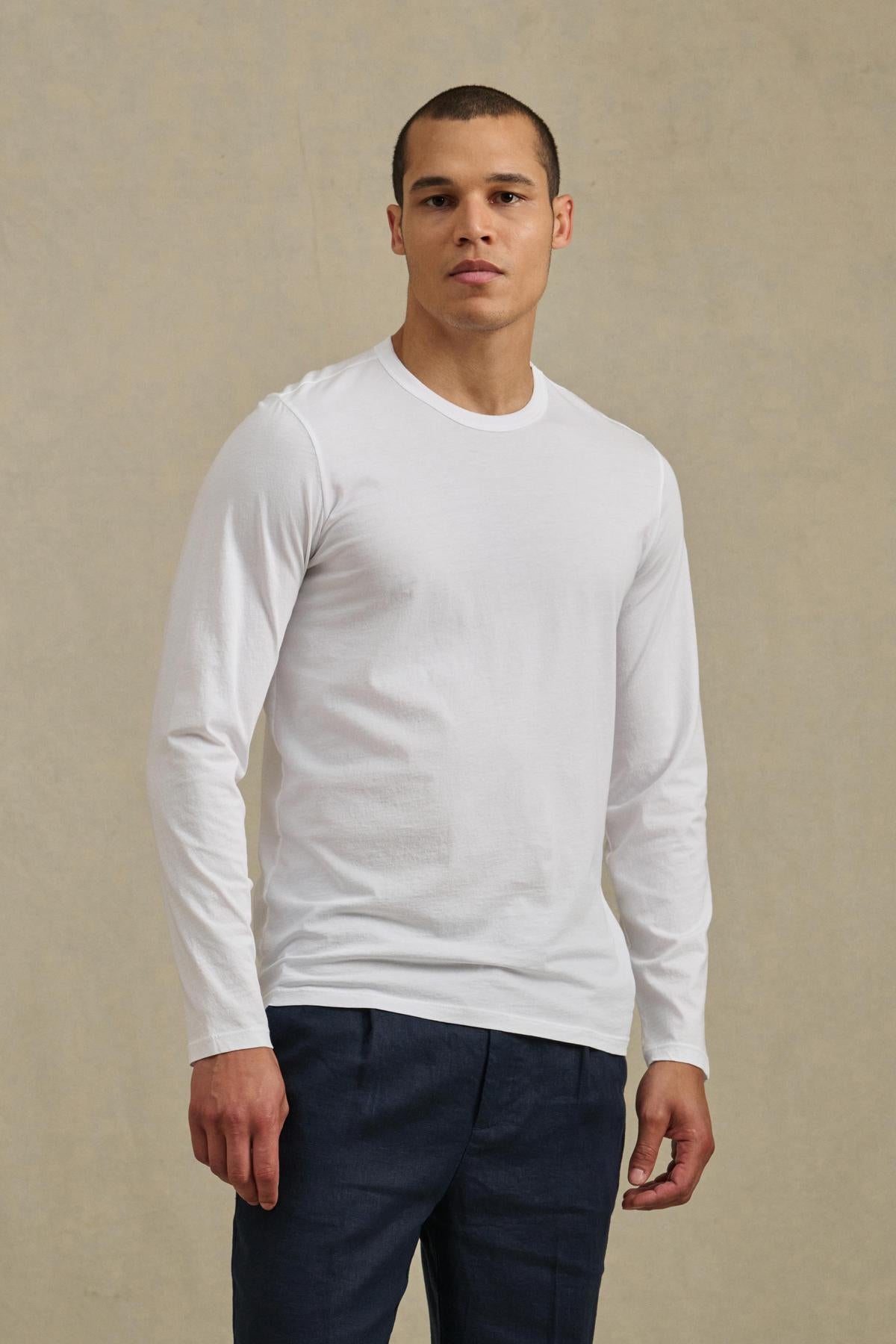 A person stands facing forward, wearing the ROWLAND TEE by Velvet by Graham & Spencer—a plain white long-sleeve shirt with a classic crew neckline—paired with dark pants against a neutral background.