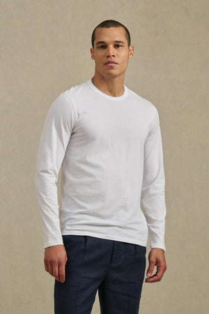 A person stands facing forward, wearing the ROWLAND TEE by Velvet by Graham & Spencer—a plain white long-sleeve shirt with a classic crew neckline—paired with dark pants against a neutral background.