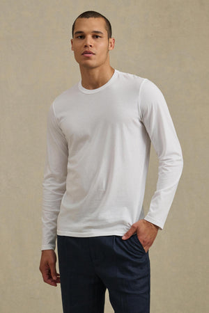 A man stands against a beige background, wearing the ROWLAND TEE by Velvet by Graham & Spencer—a plain white long-sleeve shirt featuring a classic crew neckline—paired with dark blue pants. He has one hand in his pocket and a neutral expression on his face.