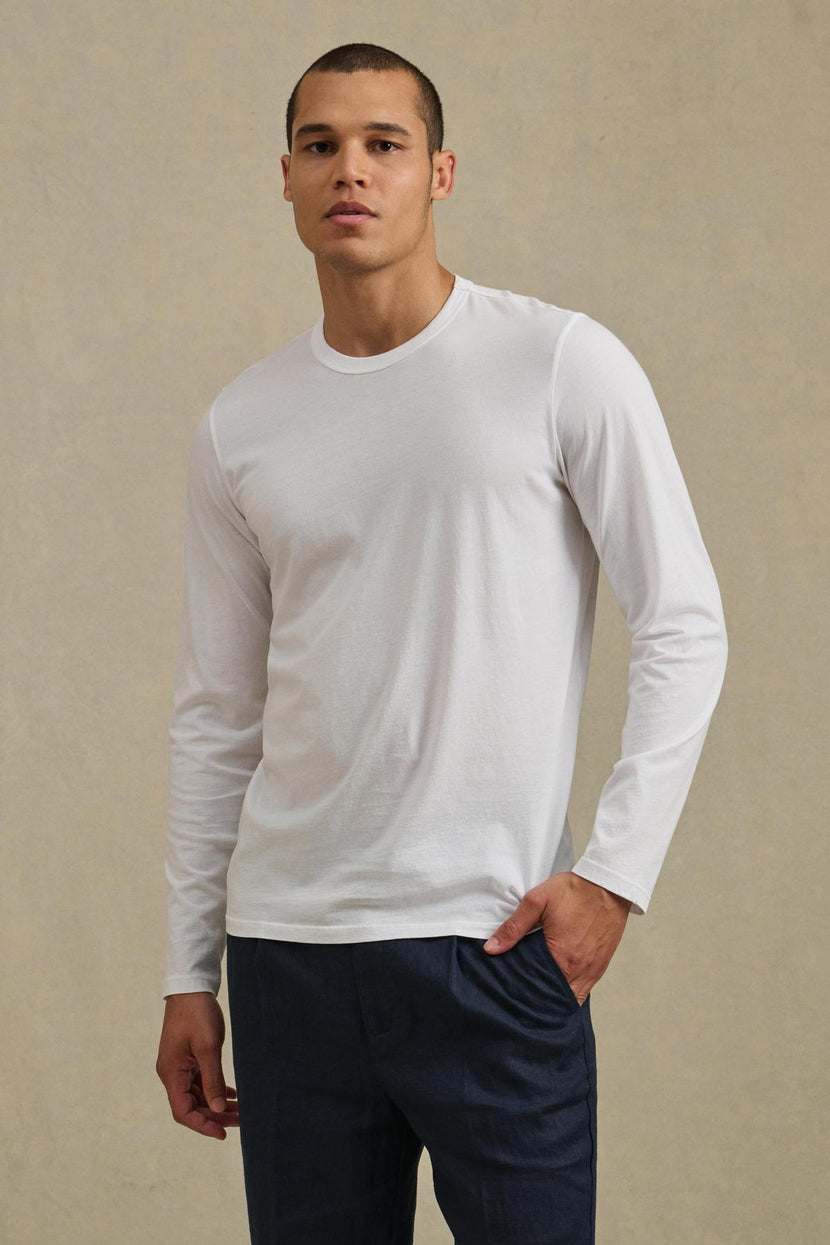 A man stands against a beige background, wearing the ROWLAND TEE by Velvet by Graham & Spencer—a plain white long-sleeve shirt featuring a classic crew neckline—paired with dark blue pants. He has one hand in his pocket and a neutral expression on his face.