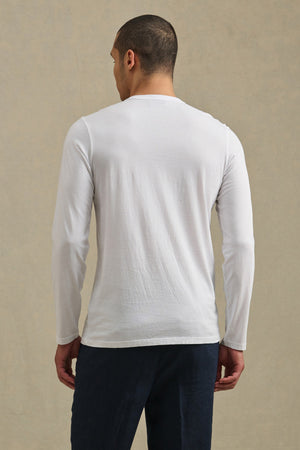 A person stands facing away from the camera, wearing the ROWLAND TEE by Velvet by Graham & Spencer—a plain white Pima jersey long-sleeve shirt with a classic crew neckline—paired with dark pants against a neutral background.