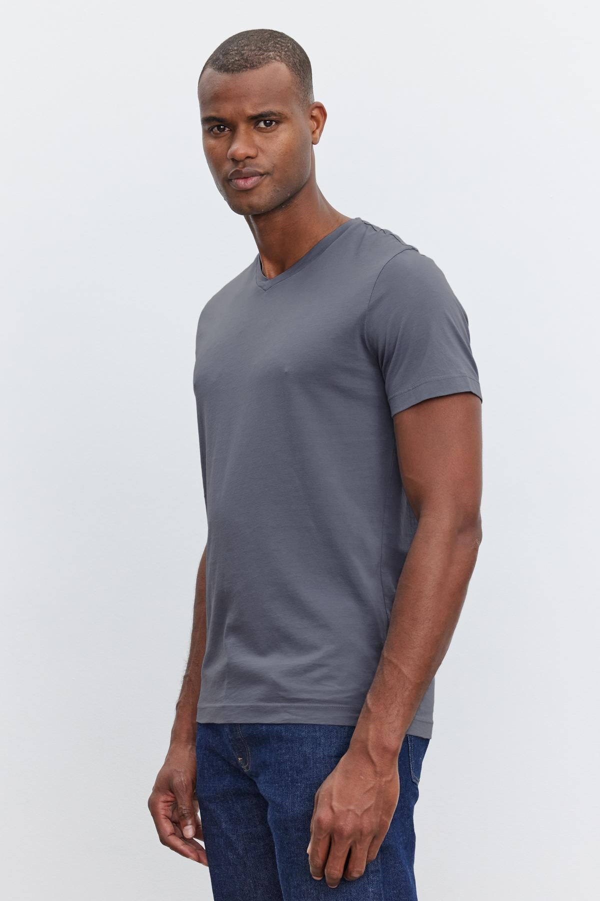   A man wearing a grey V-neckline SAMSEN TEE by Velvet by Graham & Spencer and blue jeans, made of whisper cotton knit for everyday wear, stands against a plain white background, looking at the camera with a neutral expression. 