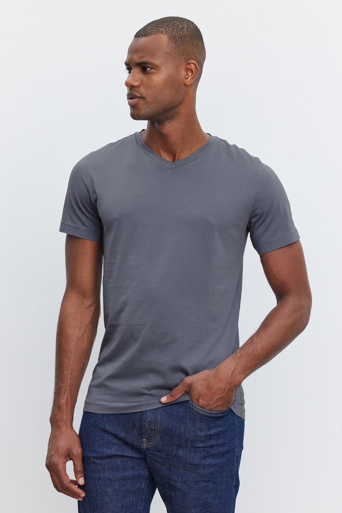 A person in a SAMSEN TEE by Velvet by Graham & Spencer with a v-neckline and blue jeans stands with one hand in their pocket, looking to the side against a plain white background—perfect for everyday wear.-37386135240897