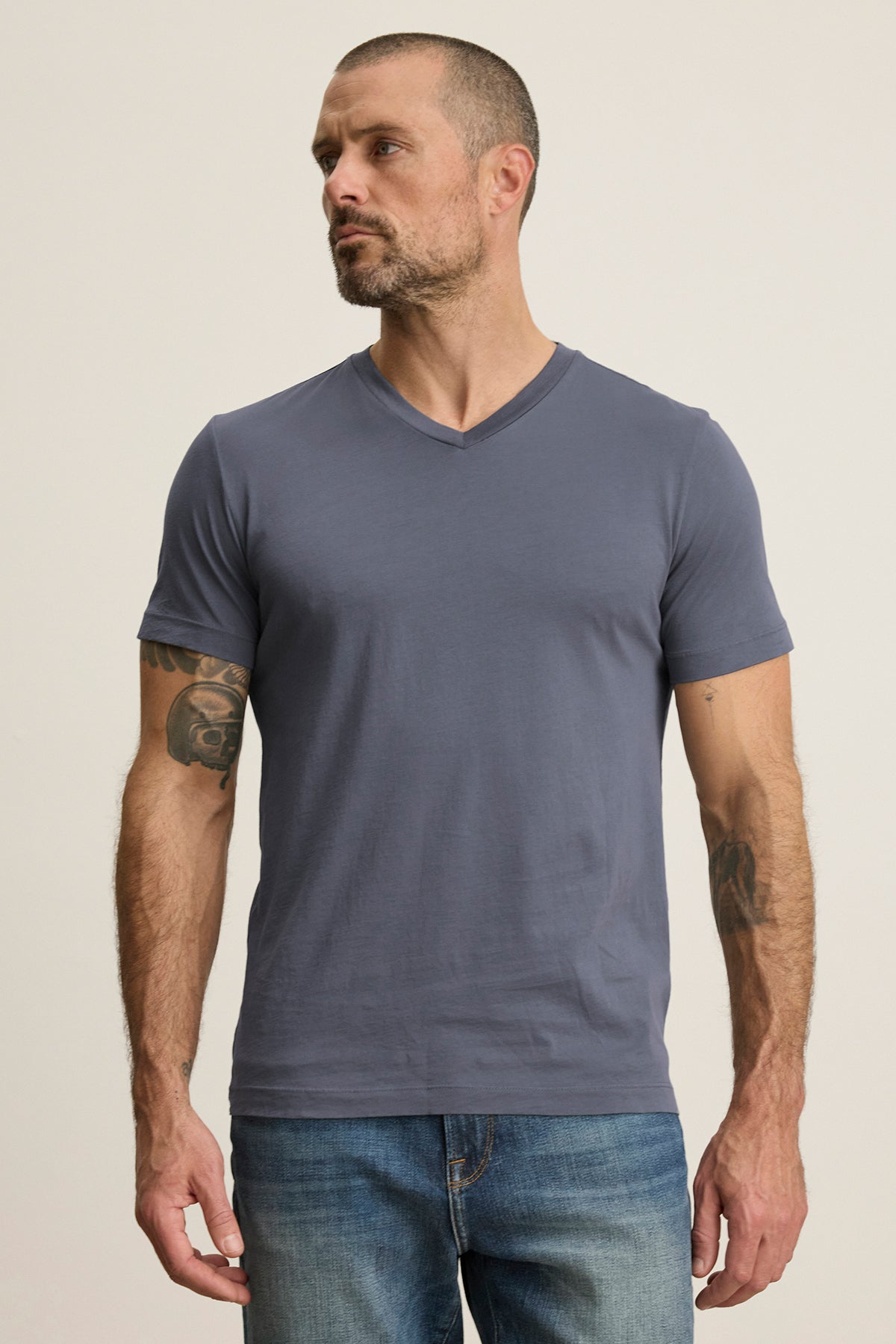   A man with short hair and tattoos stands against a plain background wearing a grey SAMSEN TEE by Velvet by Graham & Spencer, made from soft Pima jersey, paired with jeans. 