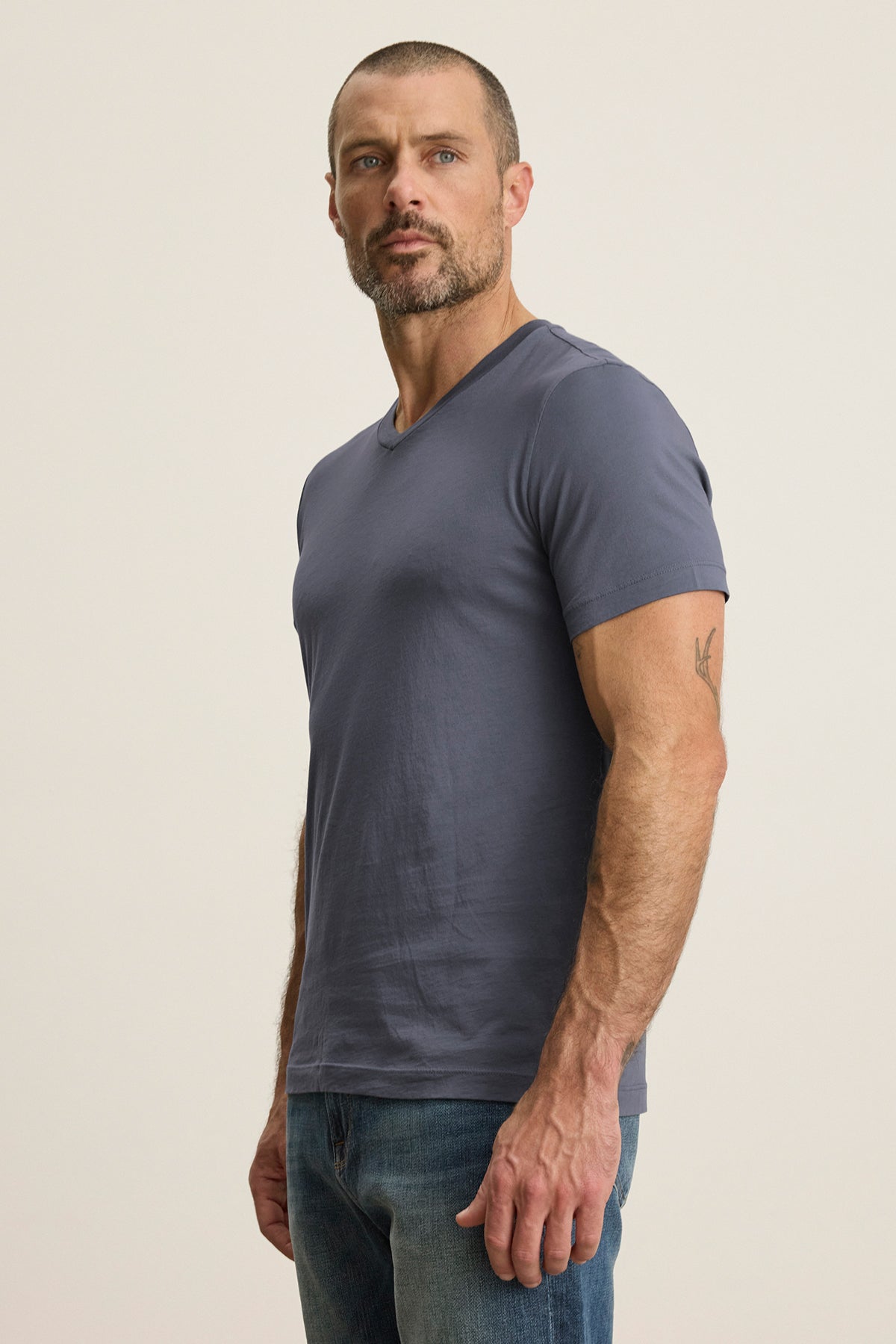   A bearded man, wearing the SAMSEN TEE by Velvet by Graham & Spencer, a gray Pima jersey V-neck T-shirt, combined with jeans, stands against a neutral background. 