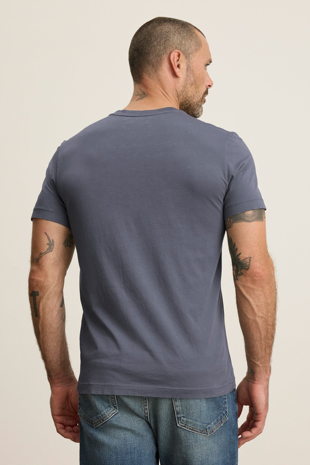   A short-haired, tattooed man in a Velvet by Graham & Spencer SAMSEN TEE—a gray Pima jersey with a subtle V-neck—pairs it with jeans as he faces away. 