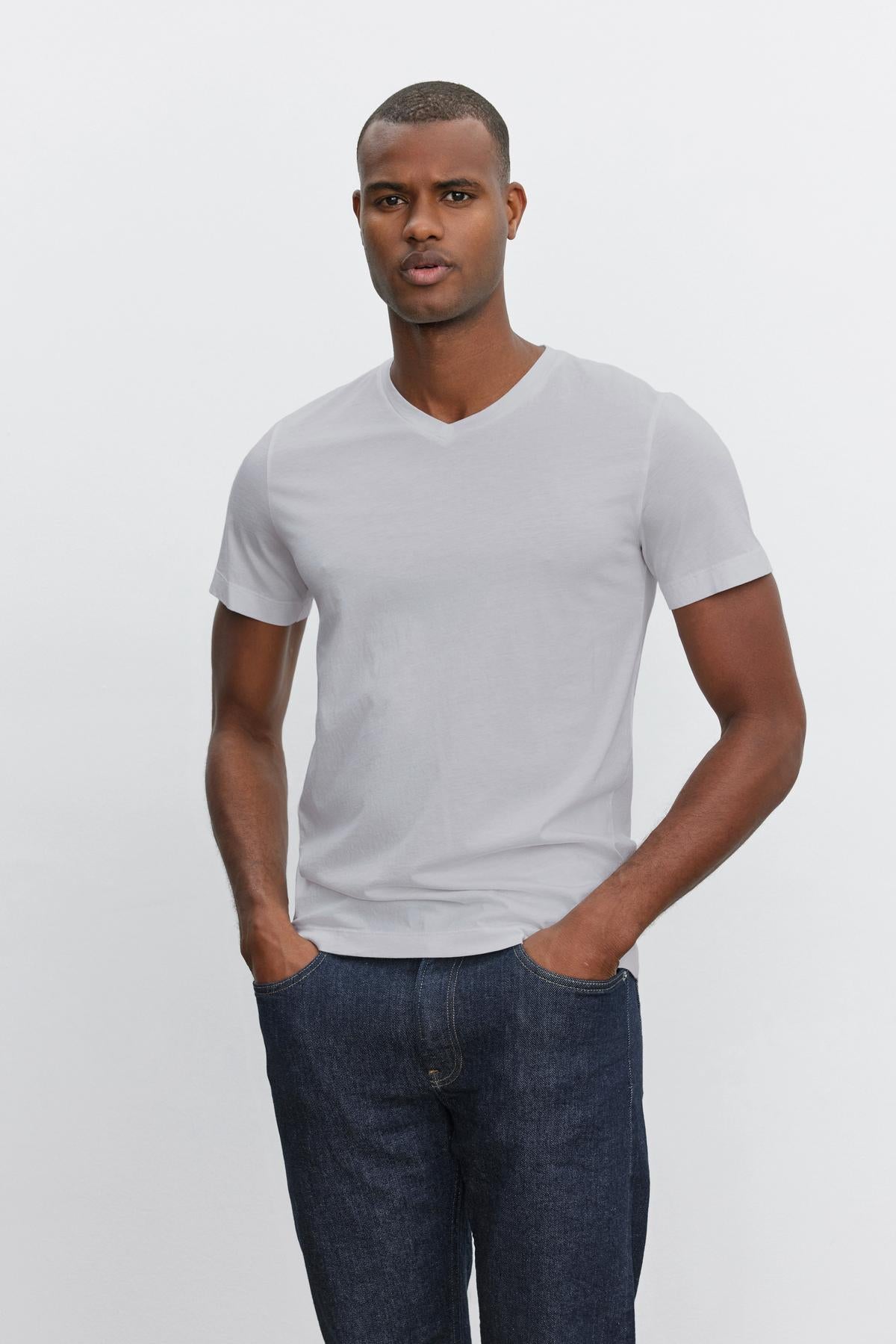 A person is standing with their hands in their pockets, wearing a plain white Velvet by Graham & Spencer SAMSEN TEE with a V-neckline and dark blue jeans—perfect for everyday wear—against a plain white background.-37386135339201