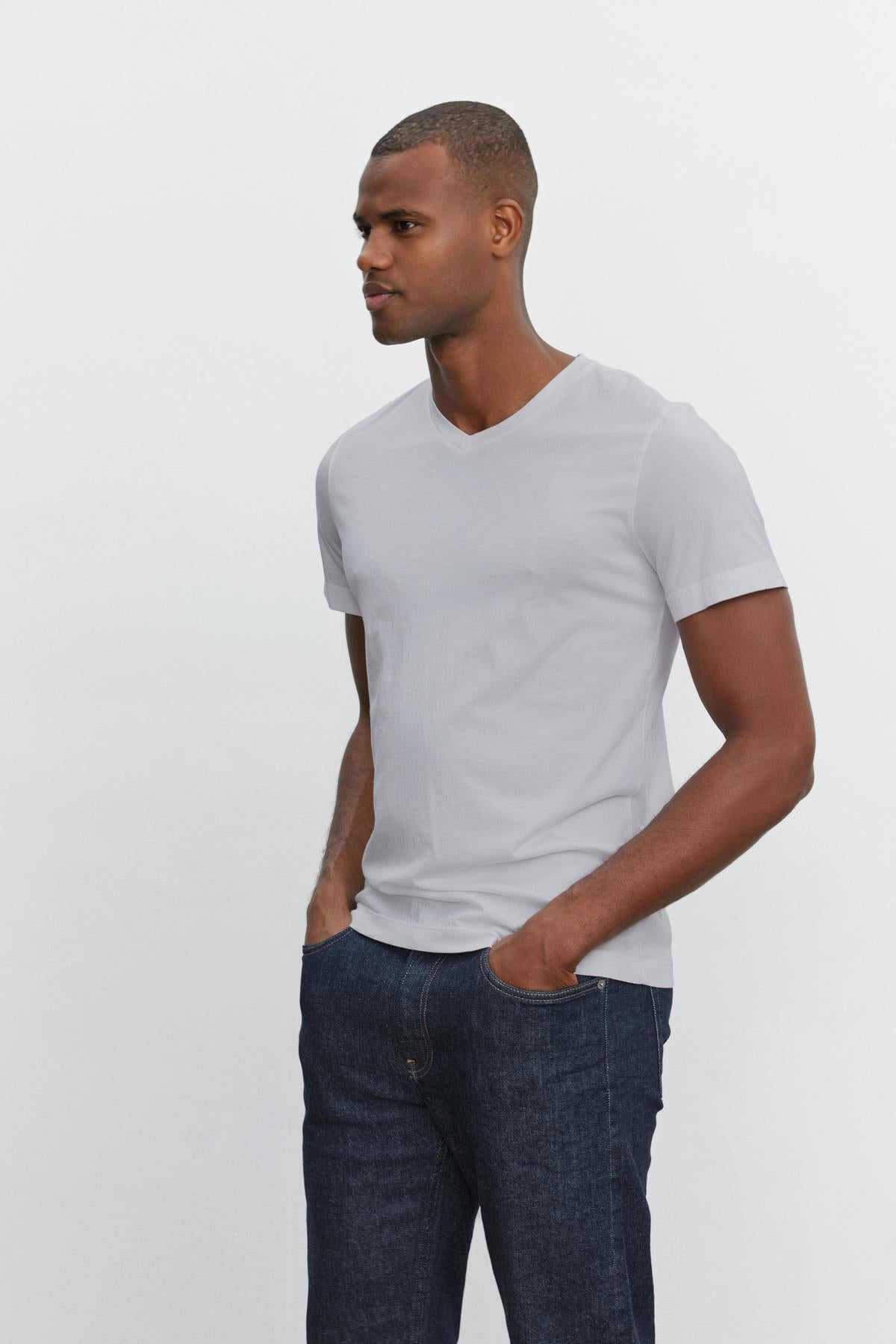   A man with short hair wears a plain light gray SAMSEN TEE made from whisper cotton knit by Velvet by Graham & Spencer and dark blue jeans. He stands and looks to the right with his hands in his pockets, against a plain white background. 