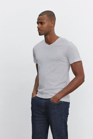 A man with short hair wears a plain light gray SAMSEN TEE made from whisper cotton knit by Velvet by Graham & Spencer and dark blue jeans. He stands and looks to the right with his hands in his pockets, against a plain white background.