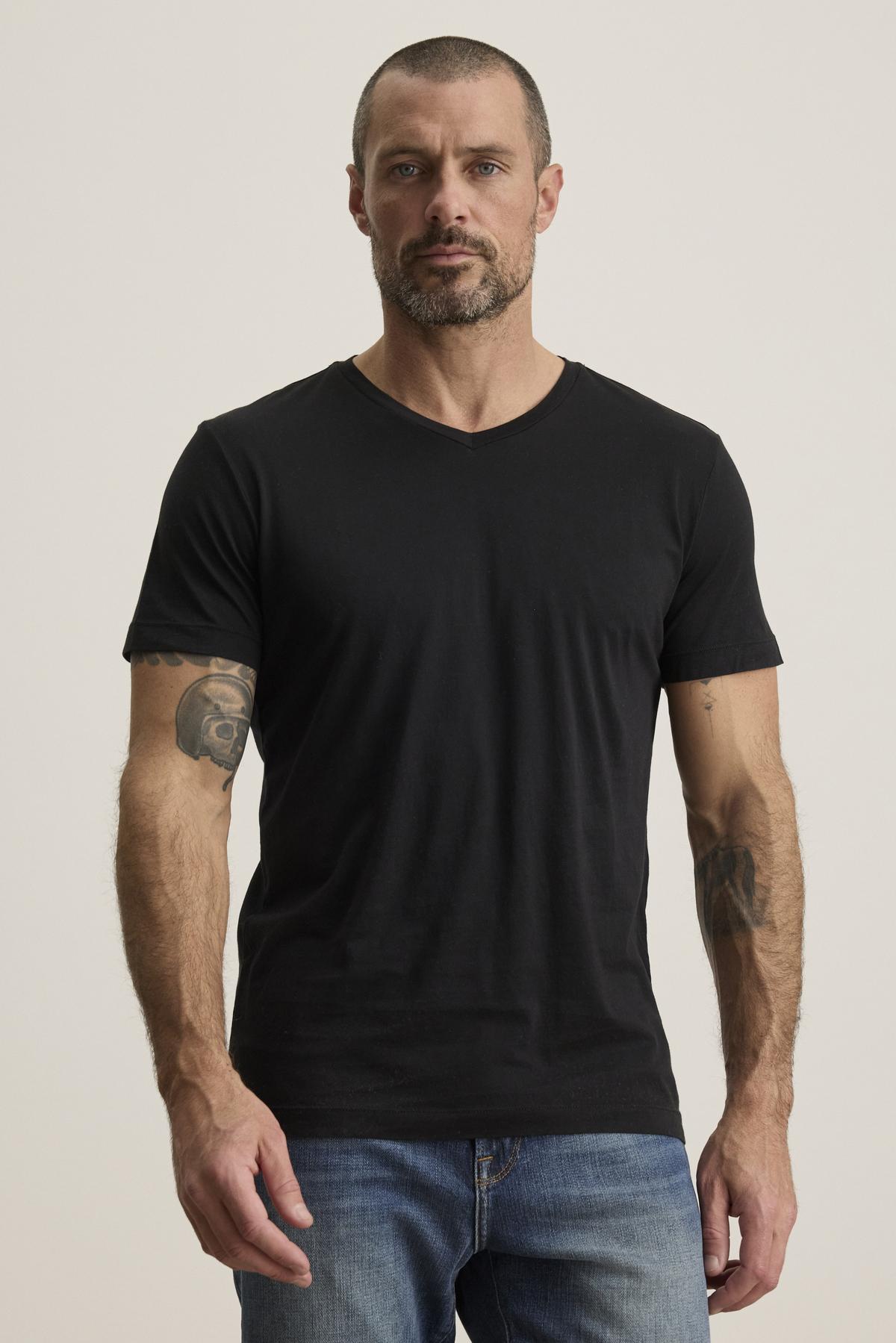   A man with short hair and tattoos on both arms is wearing a SAMSEN TEE in black by Velvet by Graham & Spencer, crafted from comfortable Pima jersey, along with jeans, as he stands against a plain background. 