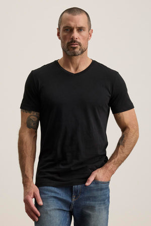 A man with short hair and a beard sports the SAMSEN TEE, a black V-neck from Velvet by Graham & Spencer, in plush Pima jersey. He pairs it with blue jeans against a plain background.