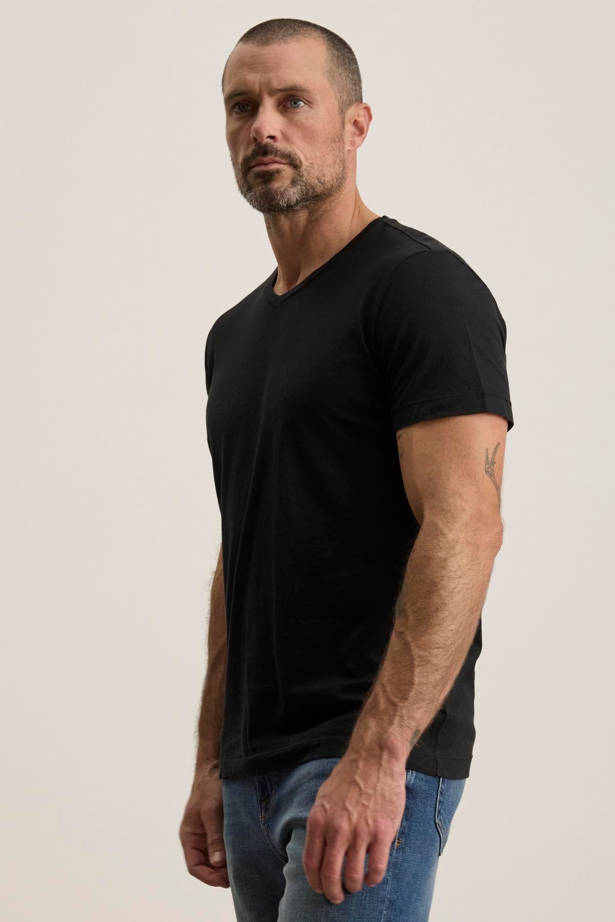 A man in a black SAMSEN TEE by Velvet by Graham & Spencer with a v-neckline and blue jeans stands against a plain background.-38504012513473