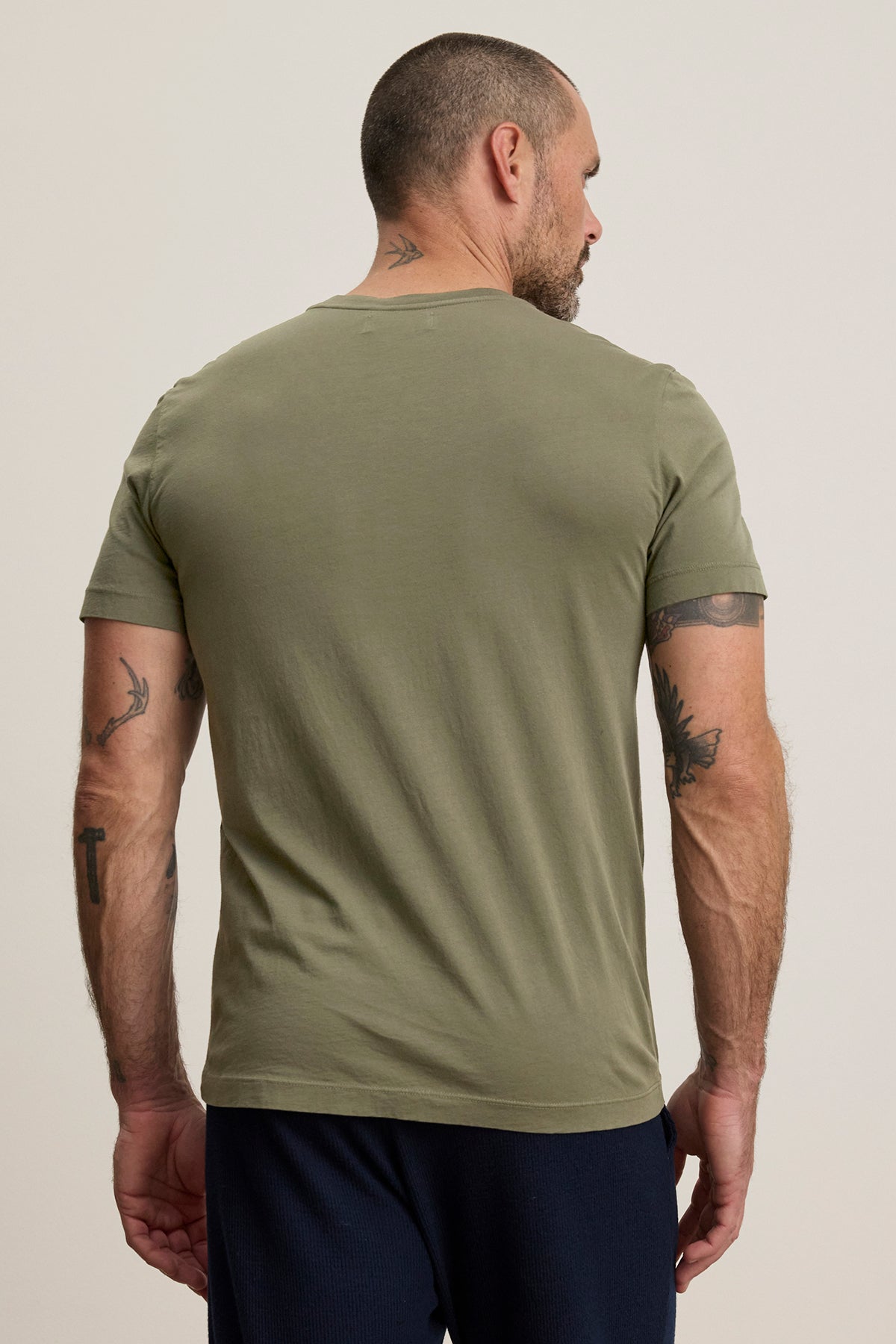   A tattooed man with a shaved head faces away from the camera, wearing Velvet by Graham & Spencer's olive SAMSEN TEE, featuring a soft Pima jersey and v-neckline, along with navy pants. 