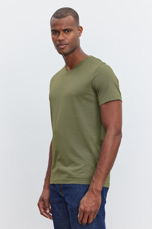 A person wearing a SAMSEN TEE from Velvet by Graham & Spencer in olive green, featuring a v-neckline, along with blue jeans, is standing against a plain background.